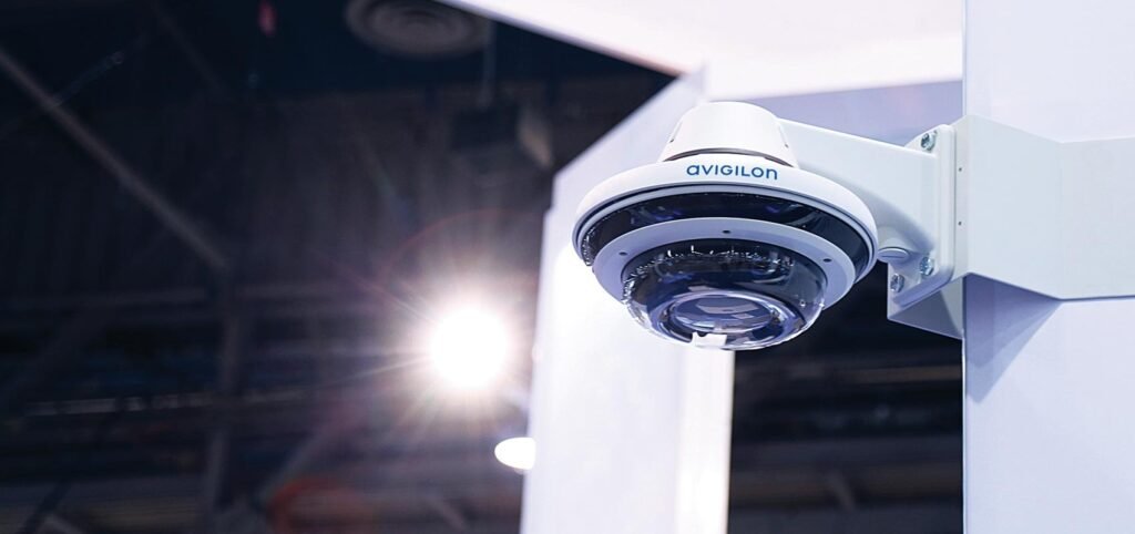 White Avigilon security camera mounted on a white pillar in an indoor exhibition hall, representing high-quality CCTV monitoring services.